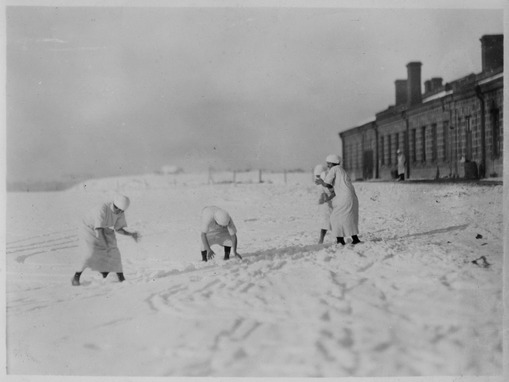 Nurses play in the snow beside a stone building.