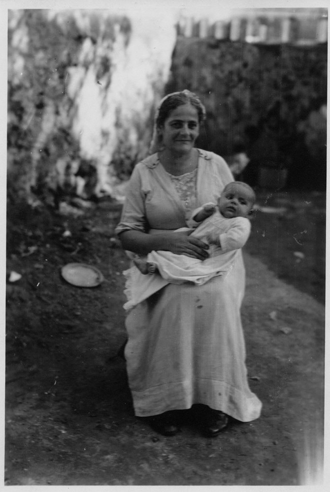 A seated woman cradles an infant in a dirt courtyard, Lebanon, c. 1921–1923.