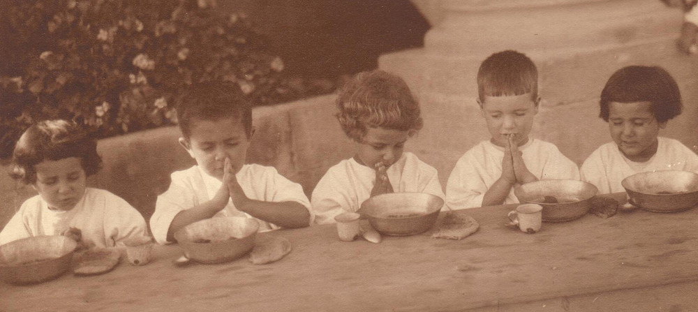 Children saying grace at the dining table of the Birds' Nest orphanage in Sidon (now Lebanon), c. 1925. Antranig Karjian appears second from the right.